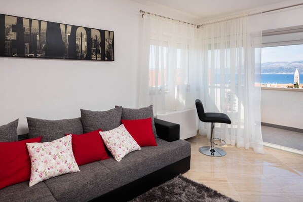 Ein modernes Wohnzimmer mit grauem Sofa, roten und floralen Kissen, einem schwarzen Sessel und einem großen Fenster mit Blick auf eine Stadtlandschaft und eine Kirche.