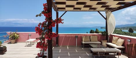 A covered outdoor seating area with a view of the sea, a pergola, and potted plants.