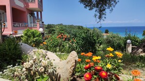A pink building with balconies, a garden with various flowers, and a view of the sea. - Studio Apartment 'GRAND  4' with Sea View, Wi-Fi and Air Conditioning (Corfu)