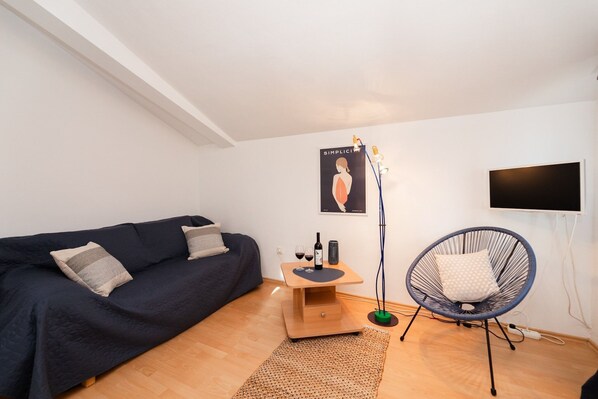 A modern living room with a dark blue sofa, a small wooden side table with a bottle and glasses, a blue chair, and a floor lamp.