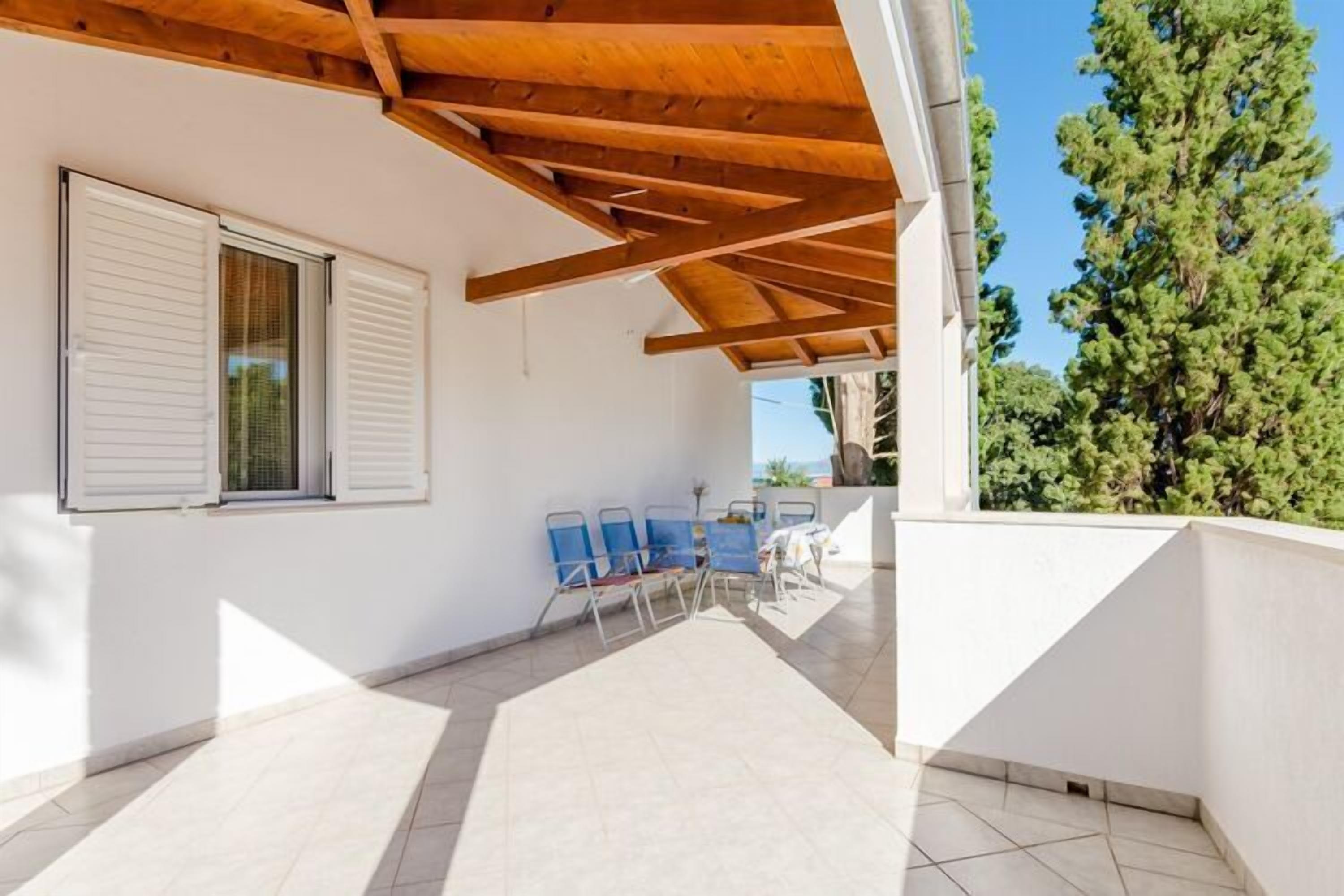 A covered outdoor area with a wooden ceiling, a white wall with a window, and a row of blue chairs.