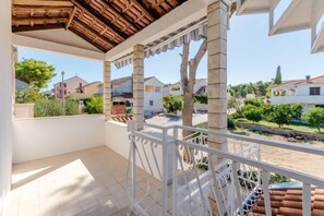 Un balcón con piso de baldosas, baranda blanca y vista a casas vecinas y follaje.