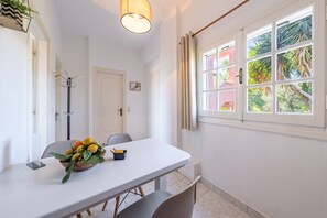 Un coin repas comprenant une table, des chaises, un panier de fruits, un porte-manteau et une fenêtre avec des rideaux.