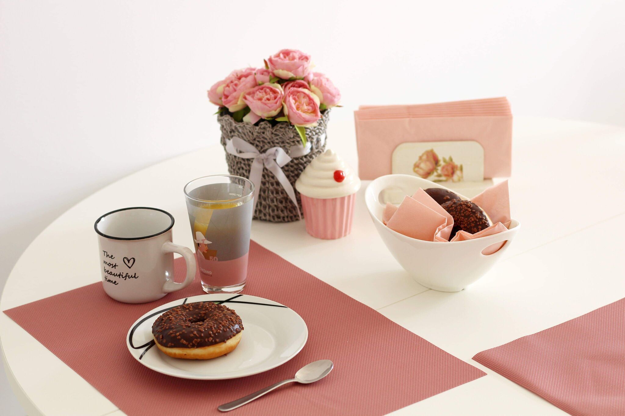 A table with a mug, a glass of juice, a cupcake, a bowl of chocolates, a plate with a donut, a spoon, a pink flower arrangement, and a card.