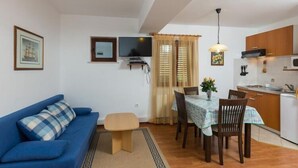 Un espace de vie compact comprenant un canapé bleu, une table à manger avec des chaises, un petit coin cuisine et un téléviseur fixé au mur.