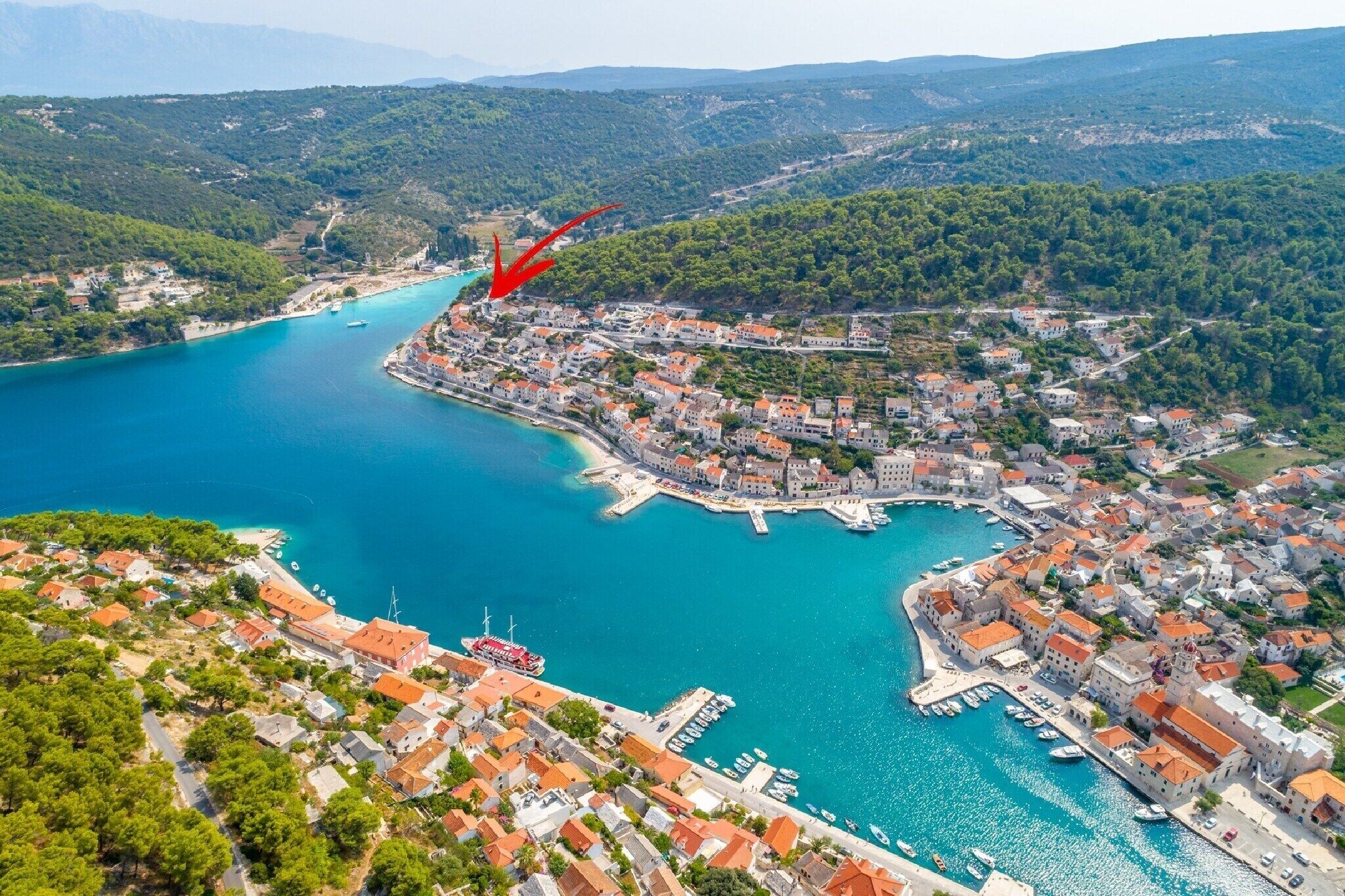 An aerial view of a coastal town with a marina, buildings with red roofs, and a clear blue sea.