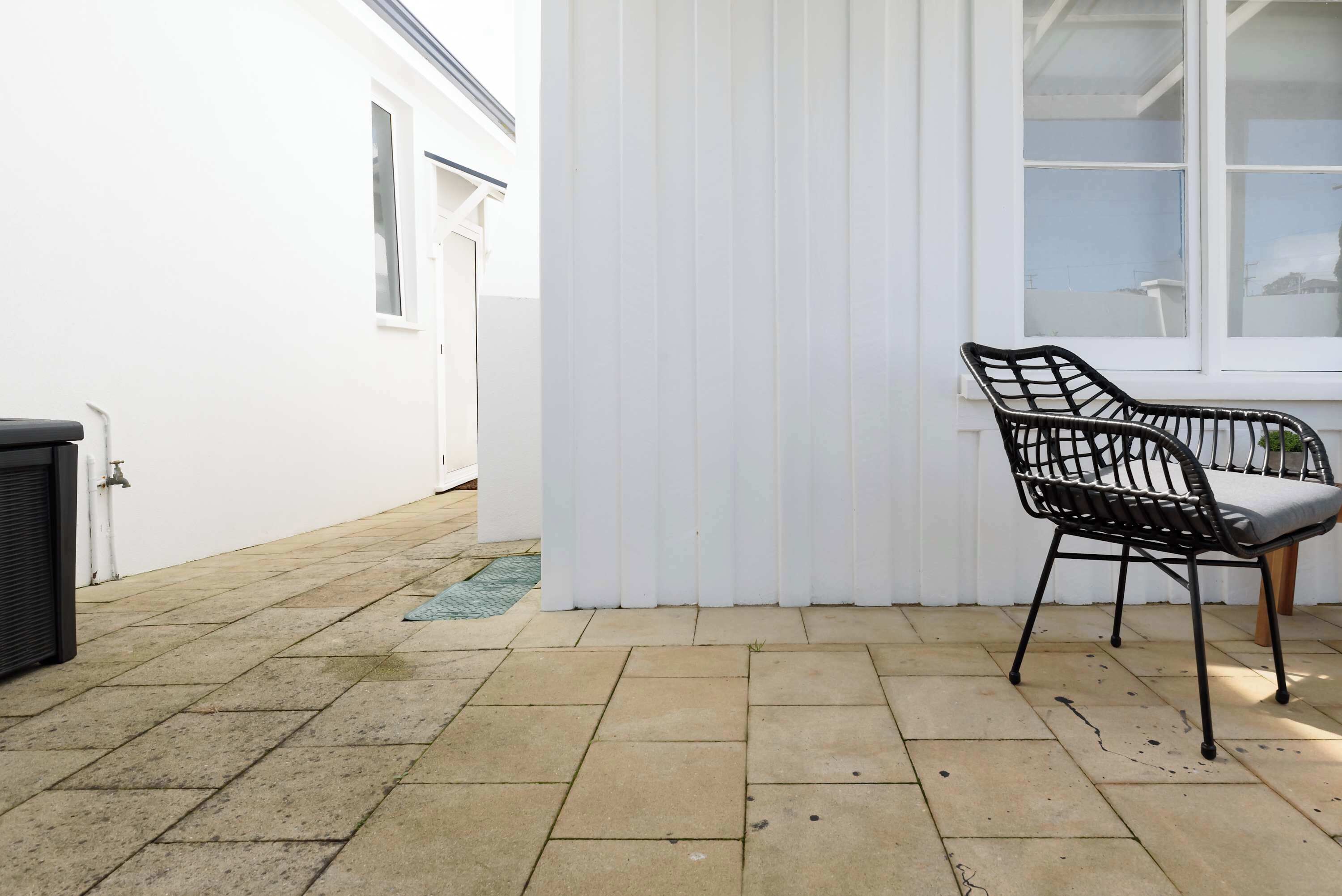 Un patio con suelo de baldosas, pared blanca, una silla de metal negro y una ventana con persianas blancas.