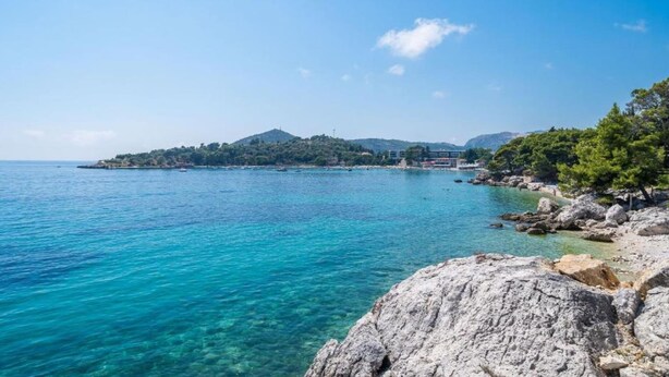 A coastal landscape with clear turquoise waters, rocky shorelines, and a distant hill with buildings.