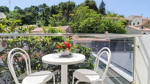 A balcony with a round table and two white chairs, a potted plant with red flowers, and a view of a town with terracotta-roofed buildings and green trees.
