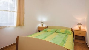 A single bed with a green and yellow bedspread, two bedside tables with lamps, a window with a sheer curtain, and a plain wall.