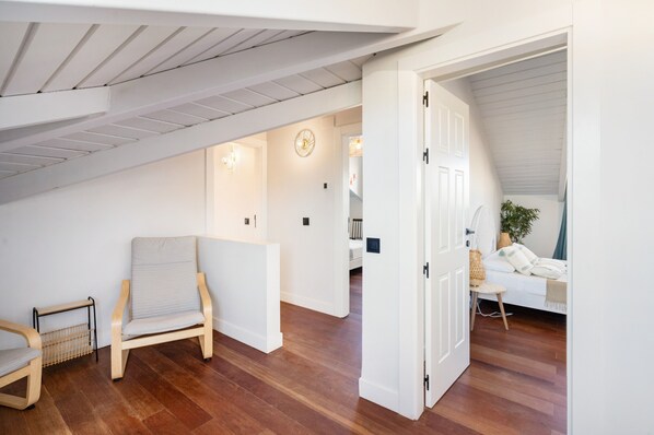 Una habitación con piso de madera, una silla, una cama con almohadas y una puerta que da a otra habitación.
