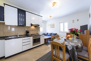 A modern kitchen with white cabinets, a stove, and a coffee maker. There is a dining area with a table set for two, a sofa, and a TV.