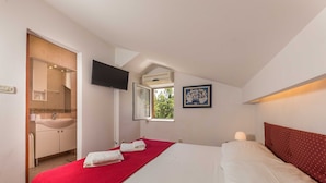 Une chambre à coucher avec un lit, une couverture rouge, un oreiller blanc, un téléviseur fixé au mur, une fenêtre donnant sur un paysage verdoyant, et une salle de bain avec un lavabo et un miroir.