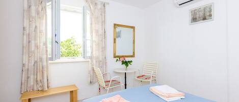 A bedroom with a bed, a bench, a small table with a chair, and an air conditioning unit.