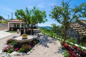 A covered outdoor seating area with stone benches and a central planter, surrounded by a gravel path and colorful flower beds. - Apartment&Rooms Savonari - Two Bedroom  Studio Apartment with Terrace (Orasac)