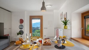A modern kitchen with a dining area, featuring a breakfast setup, a coffee machine, and a view of the outdoors. - Studio Apartment 'Maso Plocech App Vacanze' with Lake View, Private Garden and Wi-Fi (Pergine Valsugana)