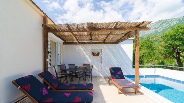 A poolside area with lounge chairs and a table, a pergola, and a view of a mountain.