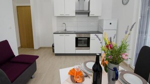 Una cocina moderna con armarios blancos, horno integrado y refrigerador. Hay una mesa de comedor con una botella de vino y un florero.