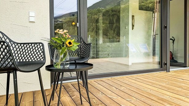 Ein moderner Außenbereich mit zwei Stühlen und einem kleinen Tisch, einer Vase mit Blumen und einer Glastür, die auf eine Terrasse mit Bergblick führt.