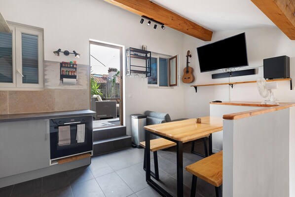 Eine kompakte Küche mit eingebautem Ofen, ein Essbereich mit Holztisch und Bänken, ein an der Wand befestigter Fernseher und eine Gitarre, die an der Wand hängt.