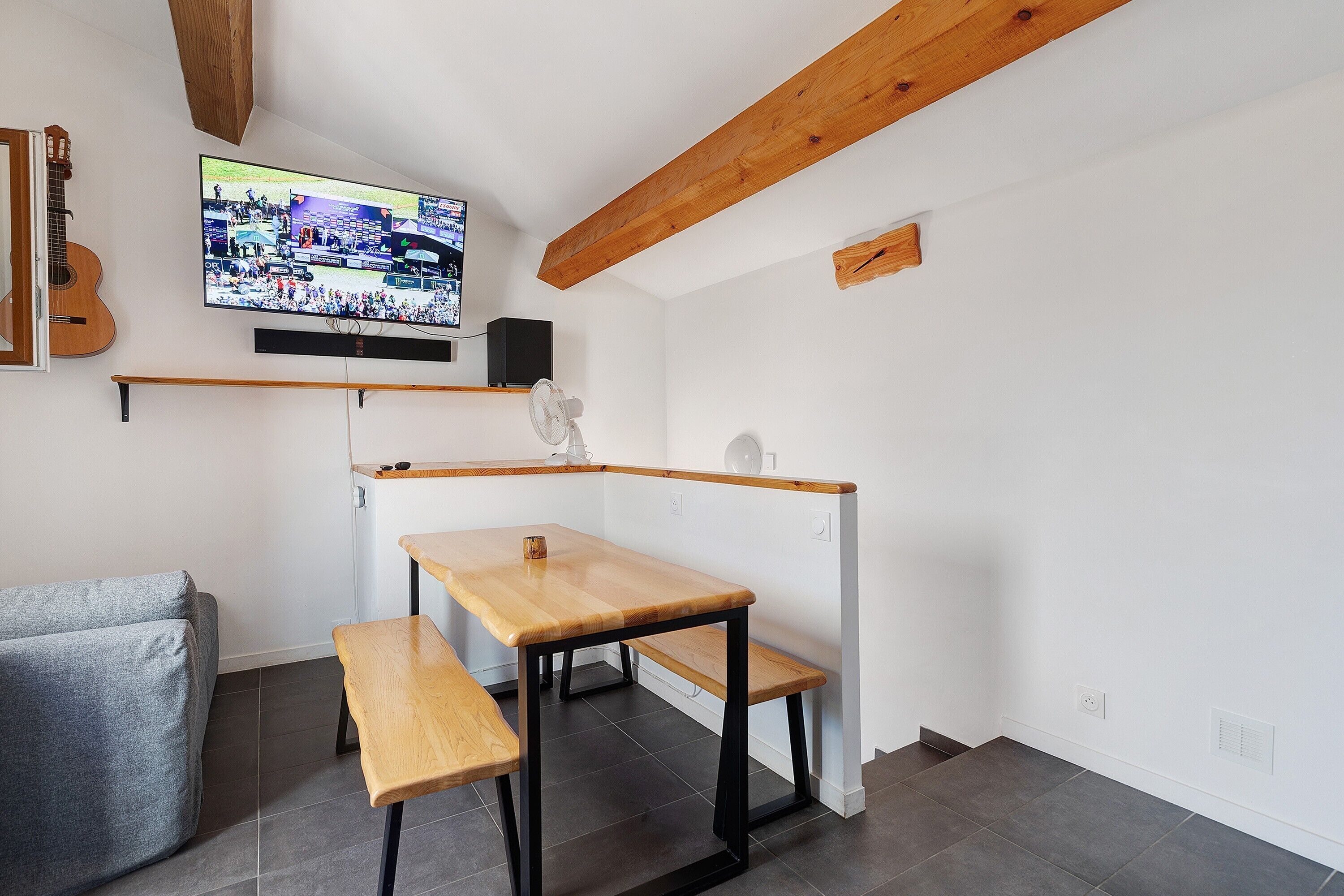 Une petite pièce avec une table et un banc en bois, un téléviseur fixé au mur et une guitare appuyée contre le mur.