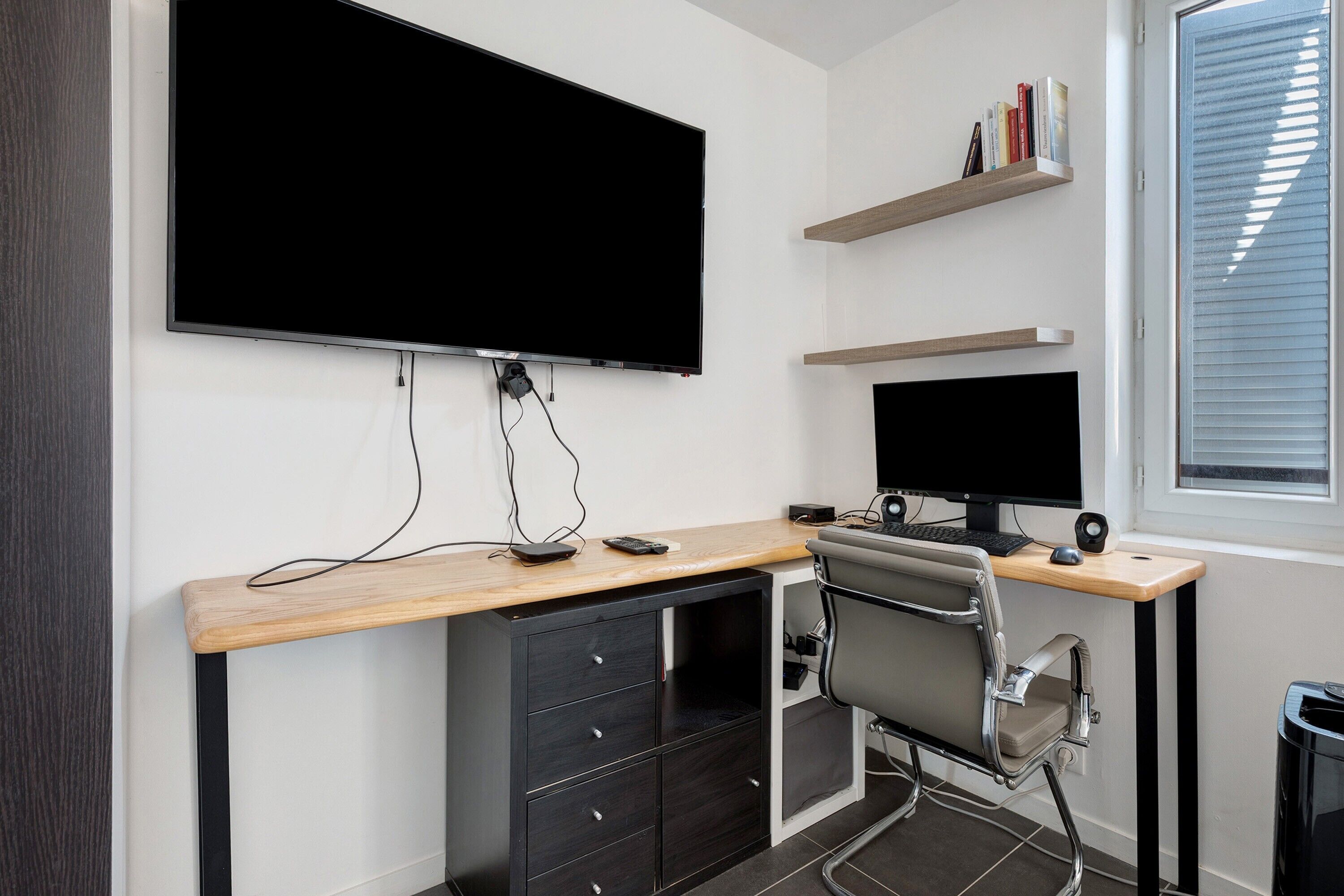 A modern office space with a large flat-screen monitor, a desk with a computer, a chair, and a window with blinds.
