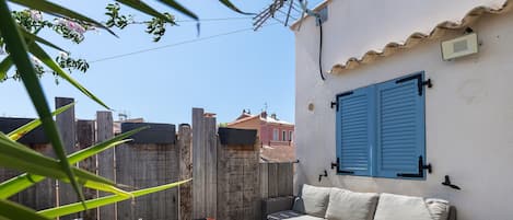 Un espacio de estar al aire libre con mobiliario de mimbre y una ventana con contraventanas azules.