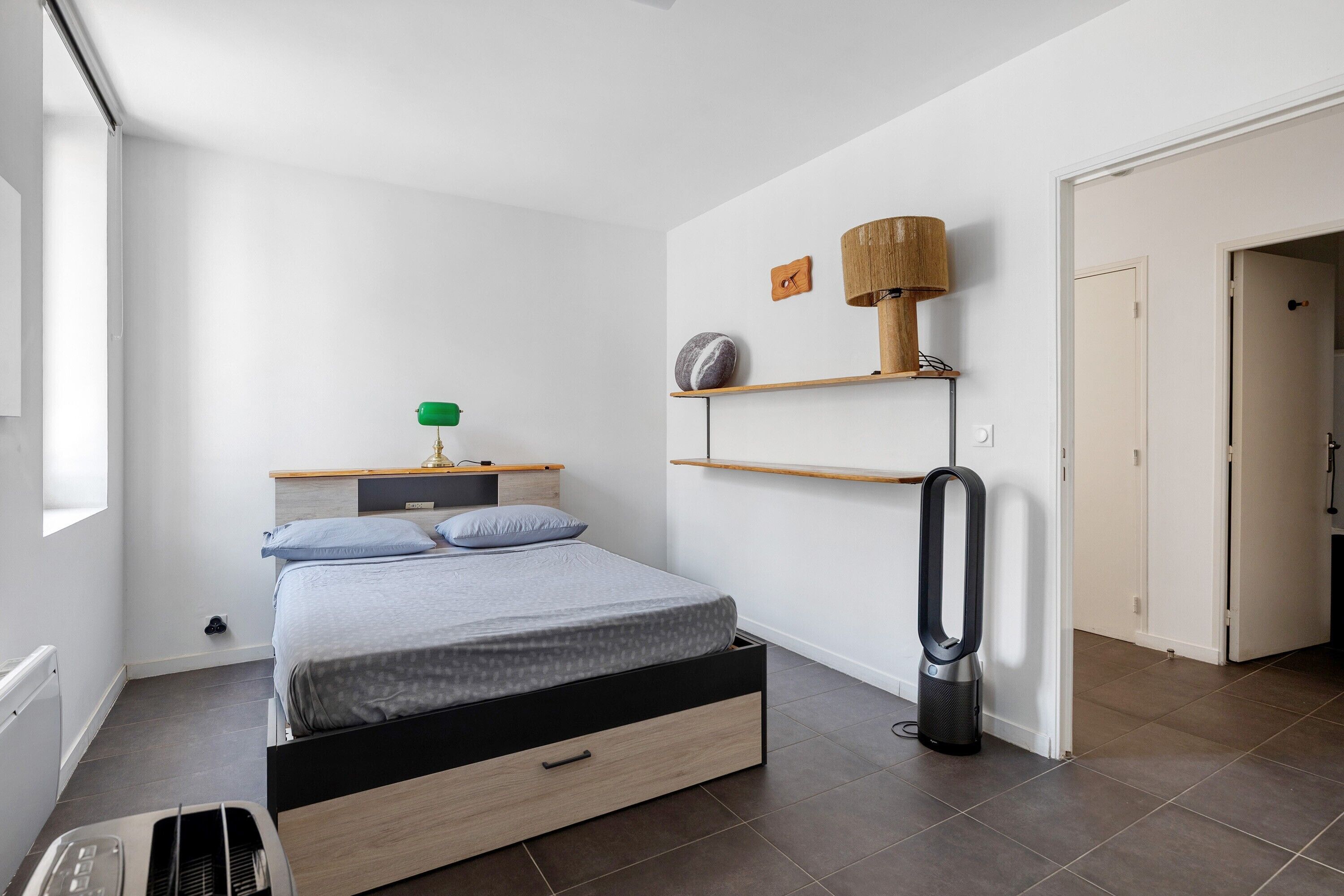 A bedroom with a bed, bedside table, lamp, and a vacuum cleaner.