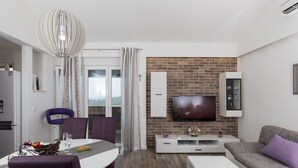 A modern living room with a TV mounted on a brick wall, a dining area with purple chairs, and a white sofa with a purple cushion.