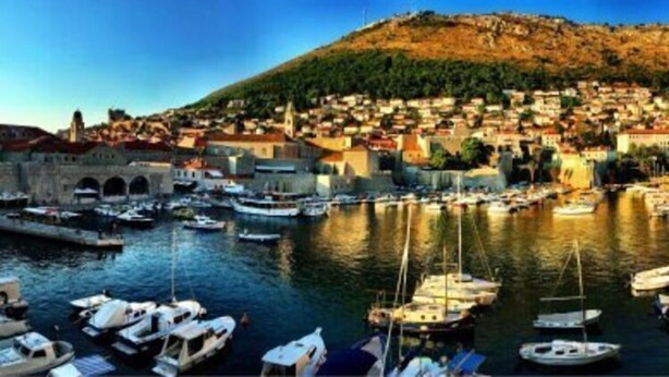 Un pueblo costero con un puerto lleno de barcos, una colina destacada con edificios, y un puente de piedra histórico.