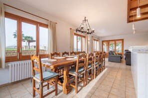 Uma sala de jantar com uma mesa de madeira comprida e cadeiras, um lustre e grandes janelas com cortinas.