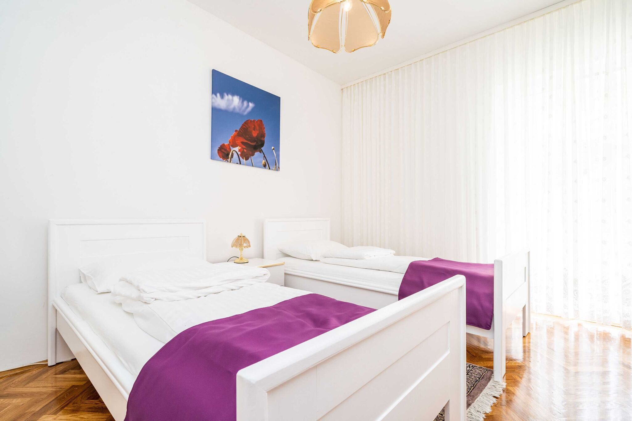 A bedroom with two beds, white bedding, a purple throw, a nightstand with a lamp, and a window with sheer curtains.