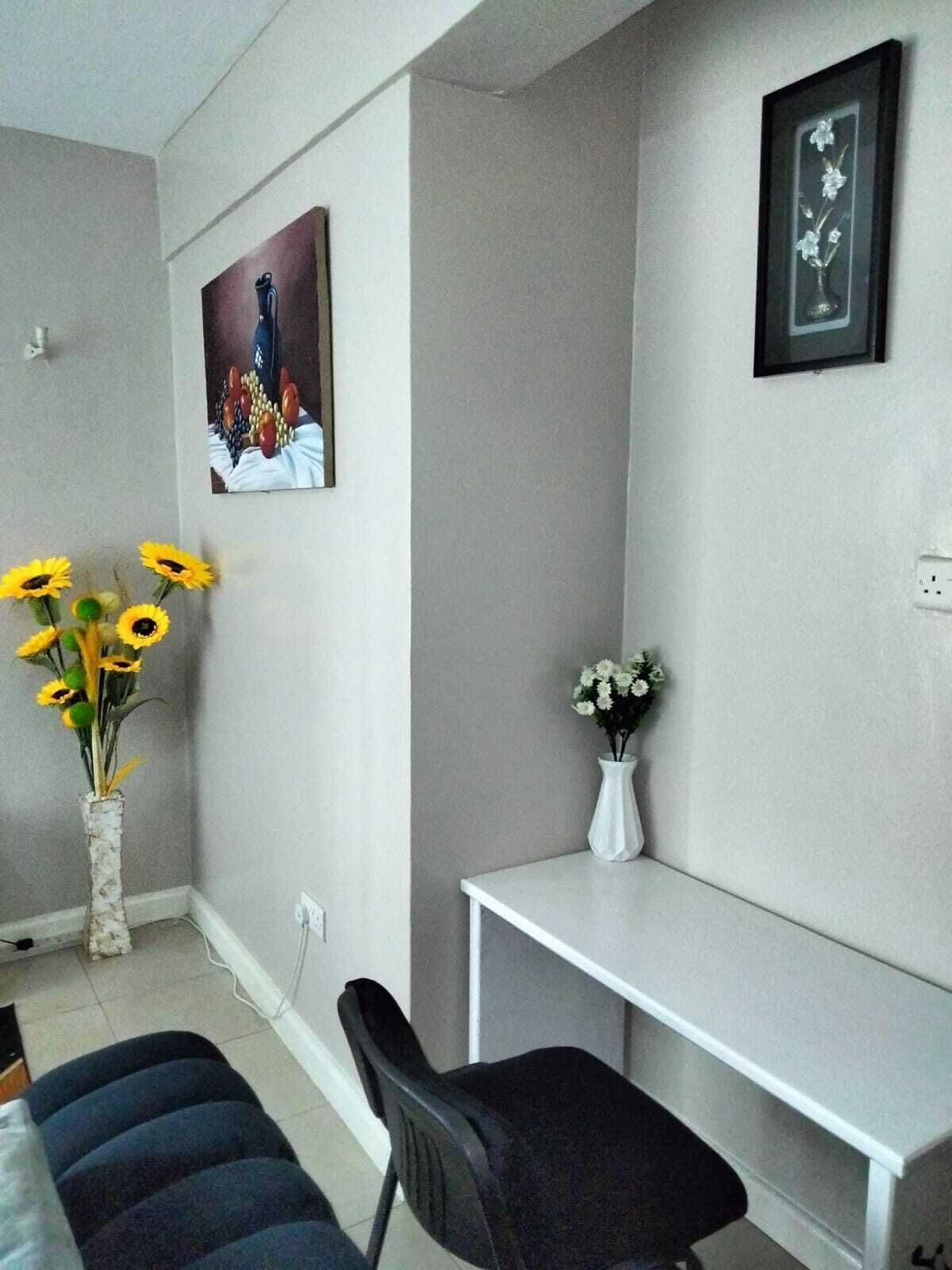 Una habitación pequeña y ordenada con una cama, un escritorio y dos sillas. Hay dos jarrones con flores sobre el escritorio.
