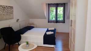 A bedroom with a bed, a chair, a small table with a plant, a window with curtains, and a painting on the wall.