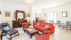A spacious living room with a red sofa, leather armchair, and a wooden coffee table. There is a dining area with a glass table and chairs, and a kitchen area with a wooden cabinet and a mirror.