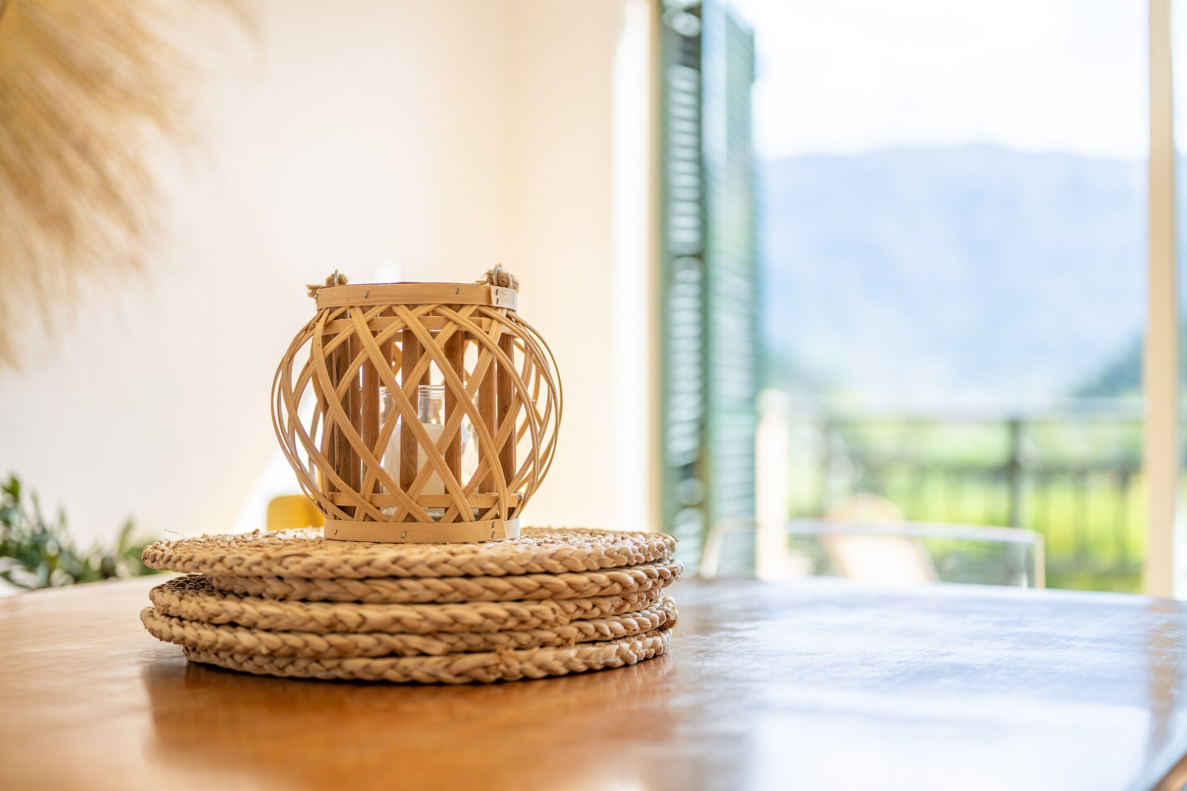 A woven coir lantern on a wooden table with a view of mountains in the background.