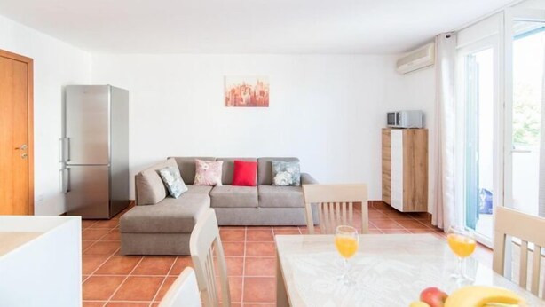 A compact living space with a sofa, a dining table with fruit and drinks, a refrigerator, and a window with a view.