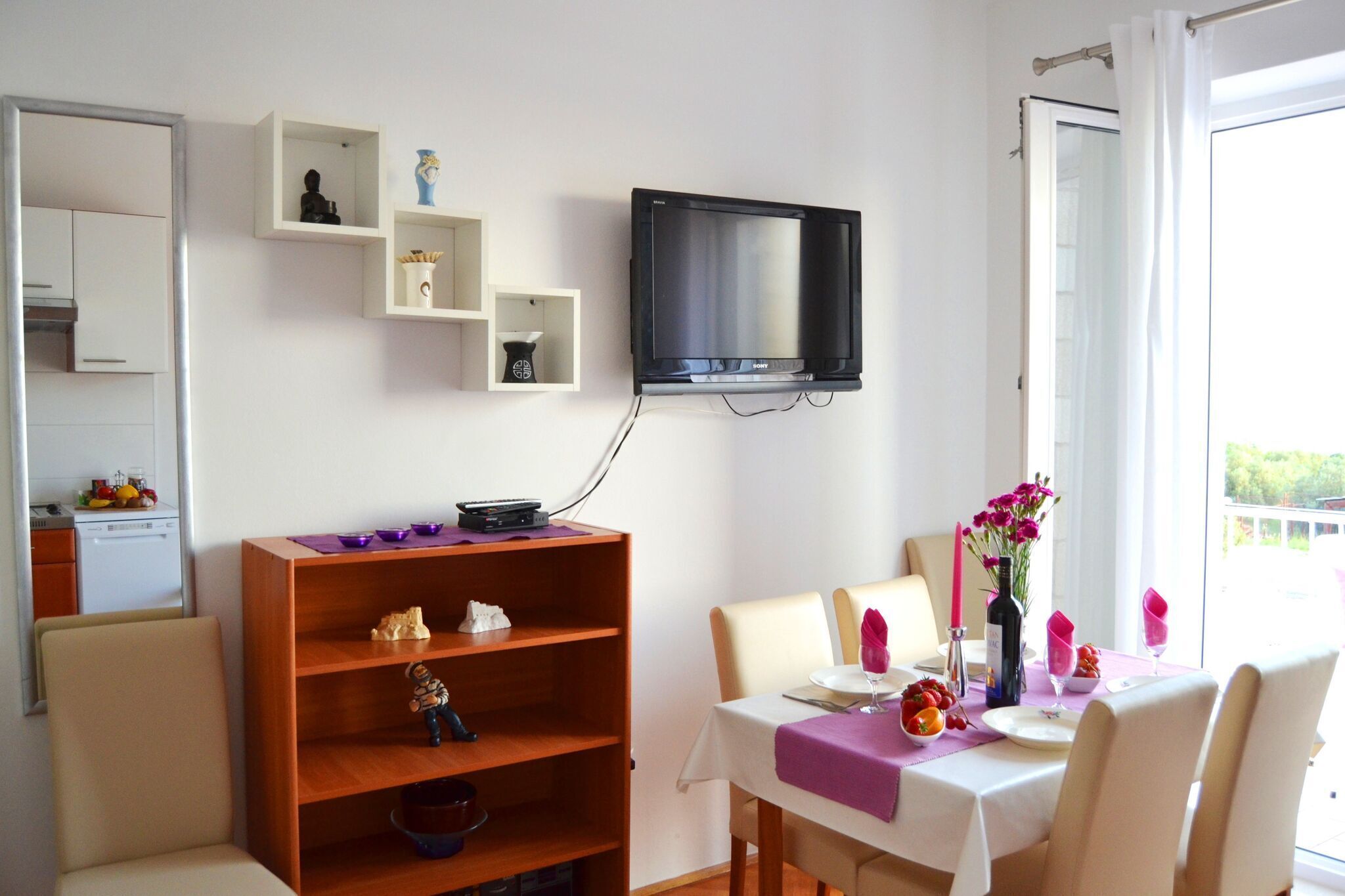 Un coin repas avec une table dressée pour deux personnes, un téléviseur fixé au mur et une étagère en bois sur laquelle sont disposés divers objets.