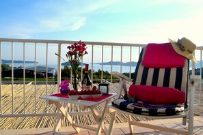 A balcony with a chair, a table with a bottle of wine, flowers, and a bowl of fruit.