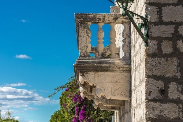 Un balcón de piedra con elaboradas esculturas y barandilla metálica, adosado a un edificio de fachada de piedra rugosa.