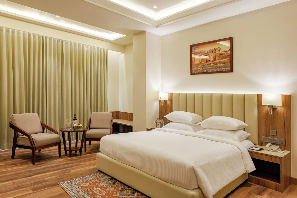 A hotel room with a large bed, two armchairs, a small table with a bottle and glasses, a nightstand with a lamp, and a painting on the wall.