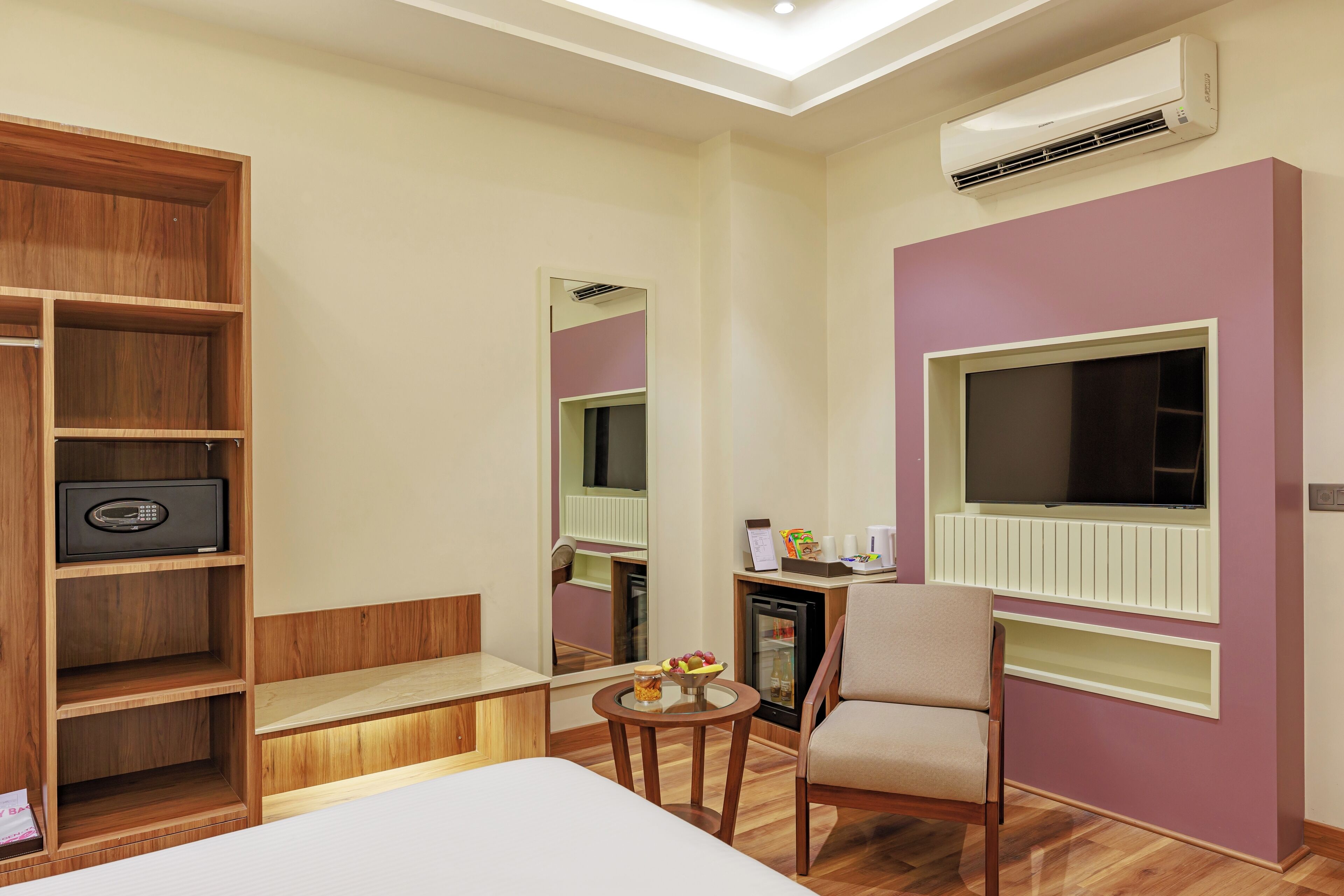 Habitación de hotel con cama, armario de madera, pequeña mesa con frutero, silla, televisor y aire acondicionado.