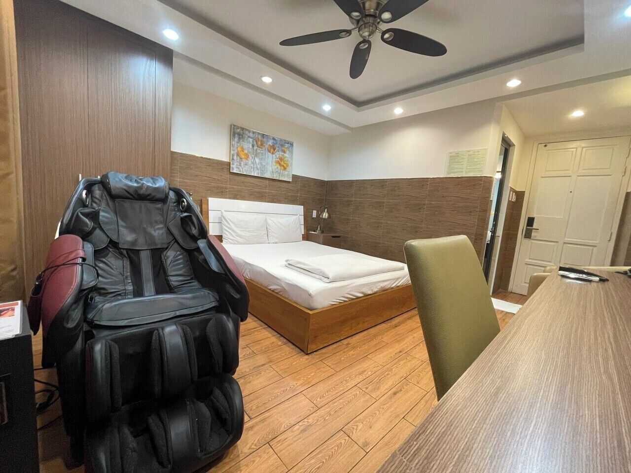 A room with a massage chair, a bed, a desk, and a ceiling fan.