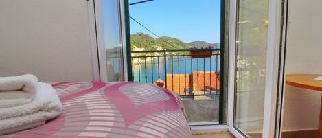 A bedroom with a bed, a window, and a view of a balcony with a railing and a wooden table.