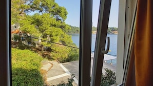 A view through a window of a coastal area with a paved path leading to a body of water, surrounded by greenery and a building with a red roof.