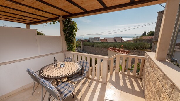 Eine überdachte Terrasse mit rundem Tisch und Stühlen, Blick aufs Meer und einer Steinmauer.