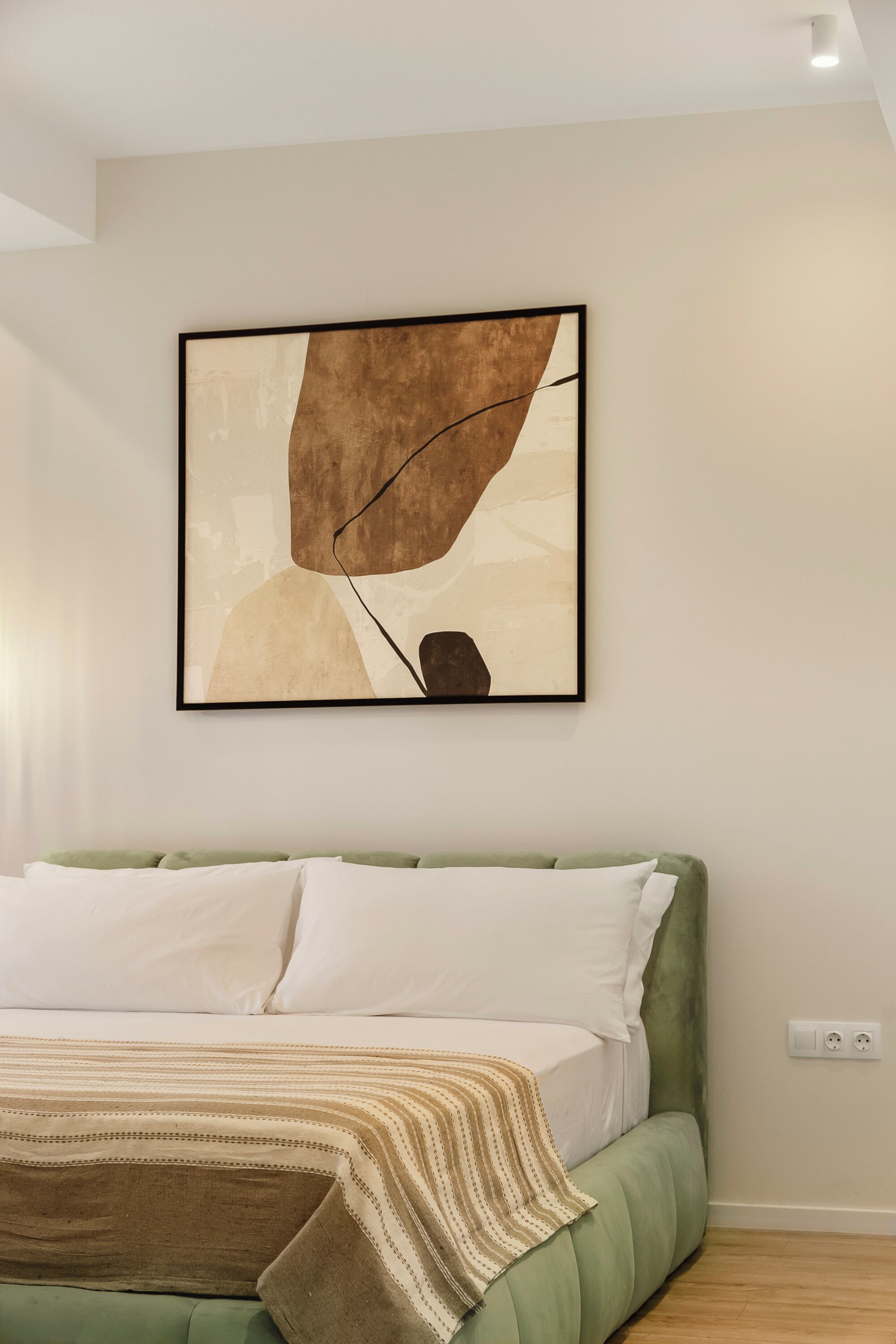 A bedroom with a green upholstered bed, white bedding, and a framed abstract painting on the wall.