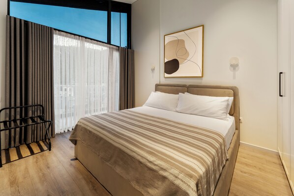 A modern bedroom with a large bed, a window with curtains, a wall-mounted abstract art piece, and a black metal chair. - Solridge Aparthotel (Tbilisi)