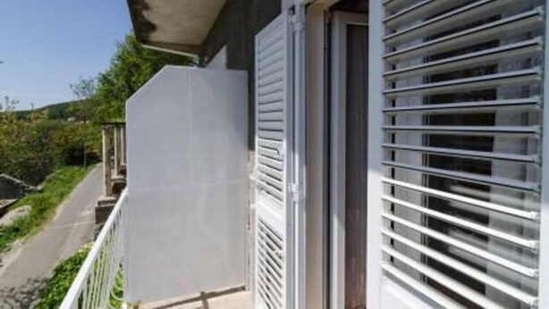 Un bâtiment doté d’un balcon, d’une fenêtre avec des stores horizontaux et offrant une vue dégagée sur la végétation environnante.