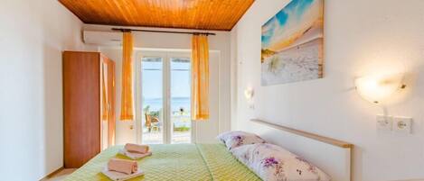 A bedroom with a wooden ceiling, a bed with a green bedspread, a painting on the wall, and a view of the sea through the window.